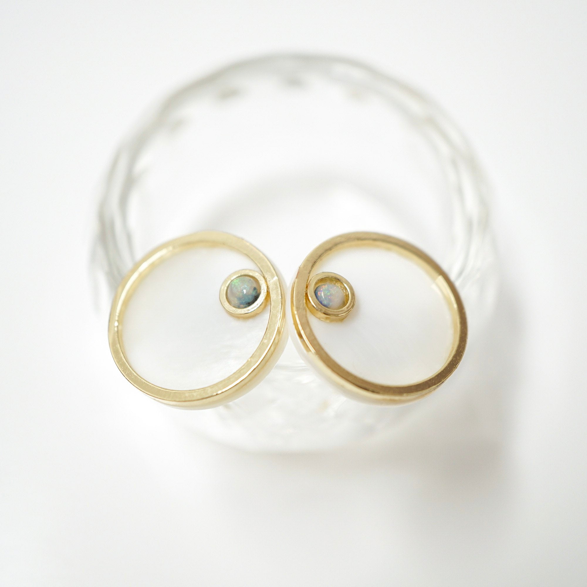 A pair of gold hoop earrings with small oval opal gemstone centers, displayed on a clear glass coaster against a white background.