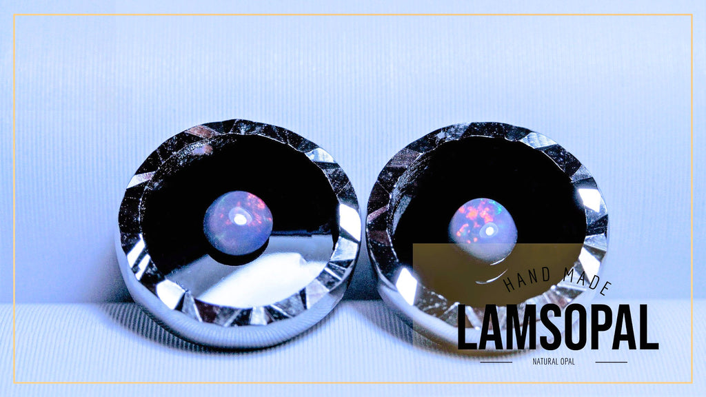 Two round Lamsopal natural opal gemstone earrings with faceted silver bezels, displayed on a light blue textured background with a gold border. A brown translucent label with 'HAND MADE LAMSOPAL' is overlaid on the right earring.
