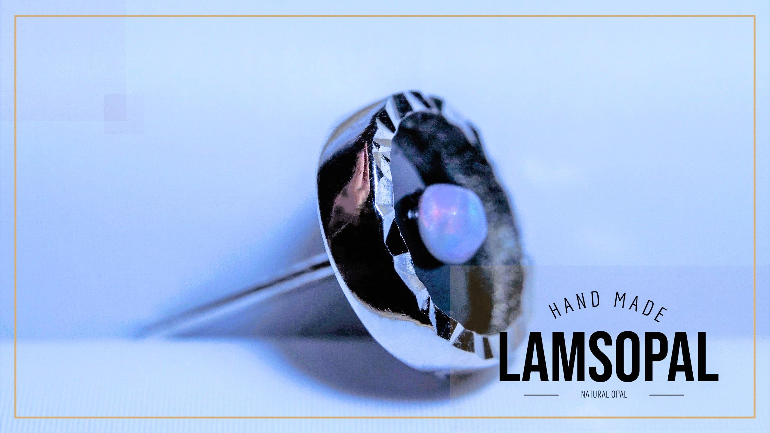 A handcrafted Lamsopal ring featuring a large, polished silver-toned dome-shaped setting with a central natural opal. The ring is shown on a light blue background with a thin gold border, and the text 'HAND MADE LAMSOPAL' is visible on the right.