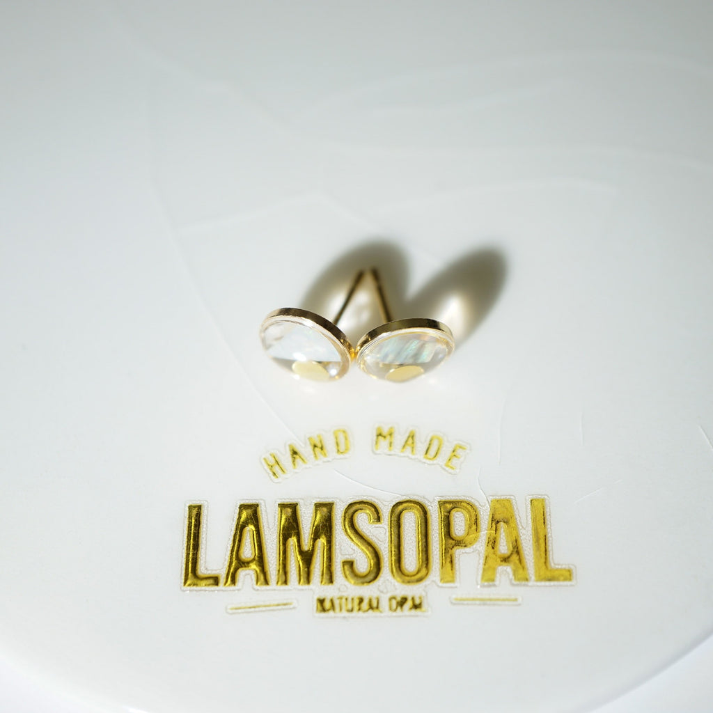A pair of gold post earrings with oval iridescent cabochon stones, displayed on a white surface above a gold foil stamp that reads 'HAND MADE LAMSOPAL NATURAL OPAL'.