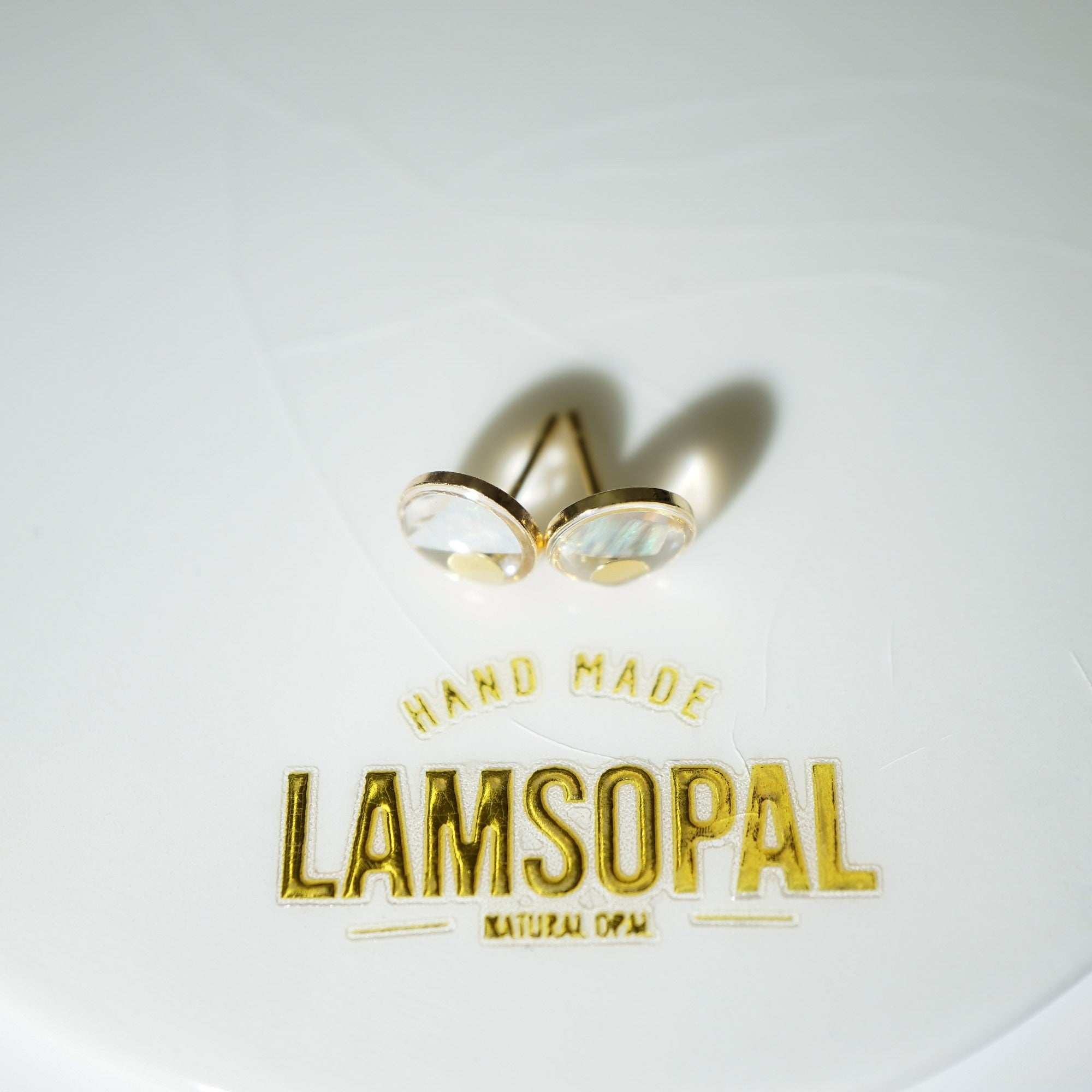 A pair of gold post earrings with oval iridescent cabochon stones, displayed on a white surface above a gold foil stamp that reads 'HAND MADE LAMSOPAL NATURAL OPAL'.