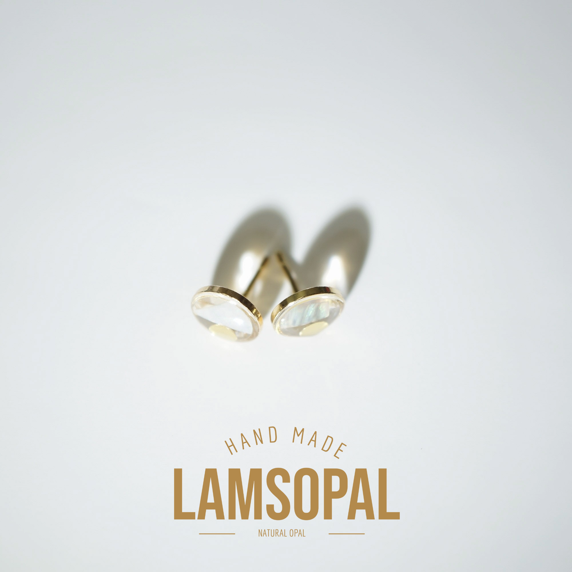 A pair of gold stud earrings with oval iridescent mother-of-pearl cabochons, displayed on a clean white background with a soft shadow. Below the earrings, the brand name 'LAMSOPAL' is printed in gold with the text 'HAND MADE' and 'NATURAL OPAL'.