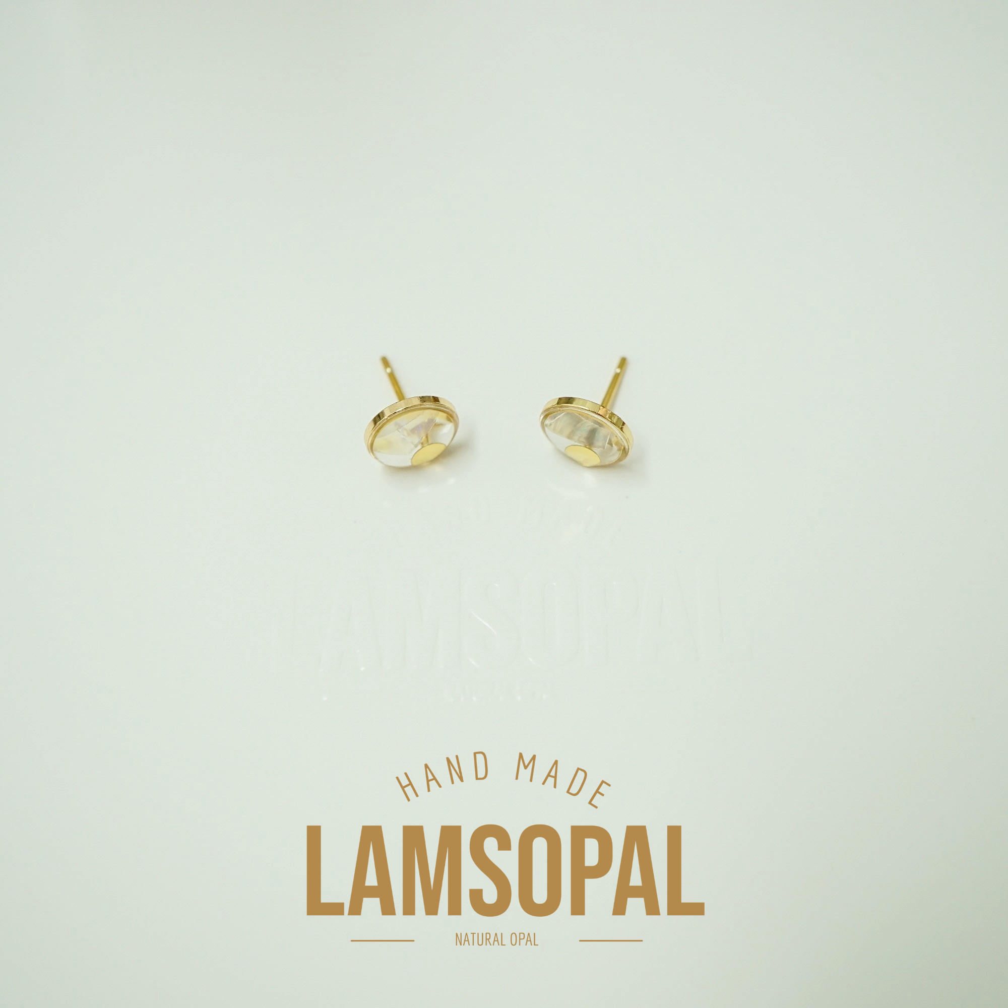 A pair of gold stud earrings with oval clear gemstones, displayed on a light cream background with a 'LAMSOPAL HAND MADE NATURAL OPAL' text overlay below.