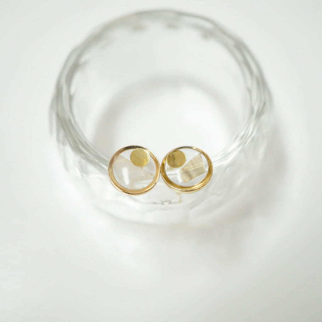A pair of gold hoop earrings with clear faceted gemstone centers, displayed on a textured clear glass stand against a white background.