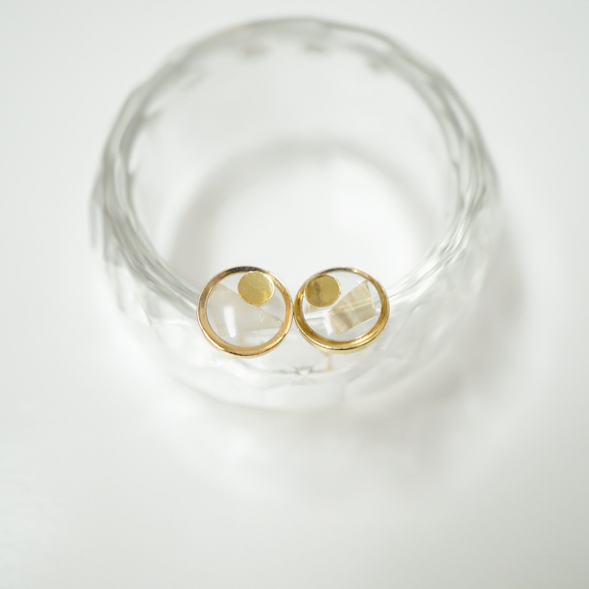 A pair of gold hoop earrings with clear faceted gemstone centers, displayed on a textured clear glass stand against a white background.