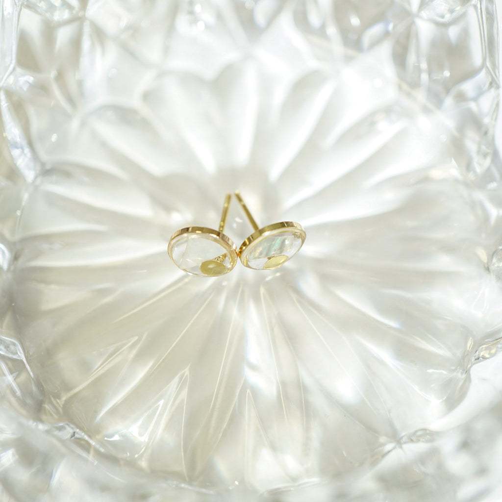 A pair of gold hoop earrings with oval clear gemstones, displayed on a textured, translucent white glass dish.