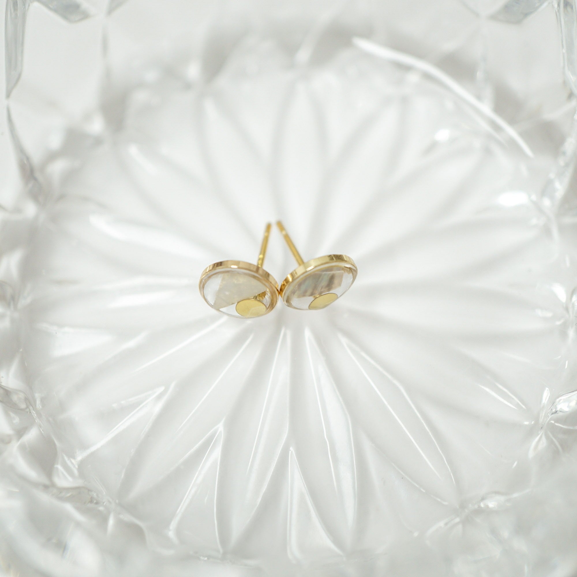 A pair of gold stud earrings with oval milky white inlays, resting on a textured white glass dish.