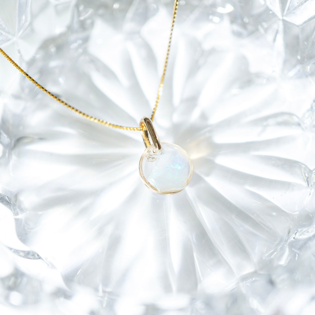 A delicate gold chain necklace with a small oval pendant featuring a milky white iridescent stone, resting on a textured white glass surface.
