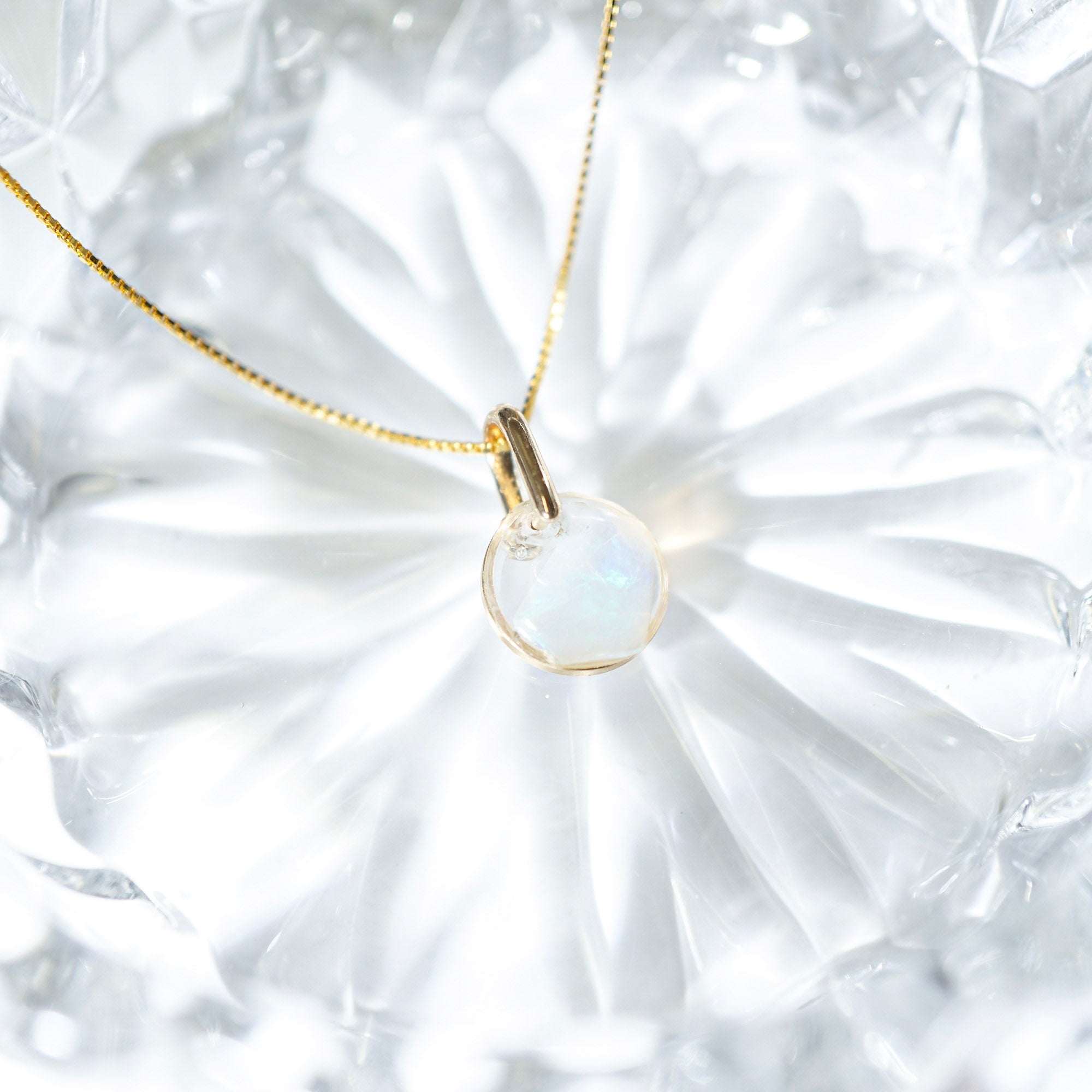 A delicate gold chain necklace with a small oval pendant featuring a milky white iridescent stone, resting on a textured white glass surface.