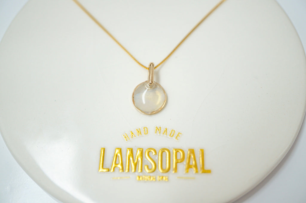 A delicate gold necklace with a small, round, clear cabochon pendant displayed on a white circular jewelry stand. The stand features gold foil text reading 'HAND MADE LAMSOPAL MUMBA'.