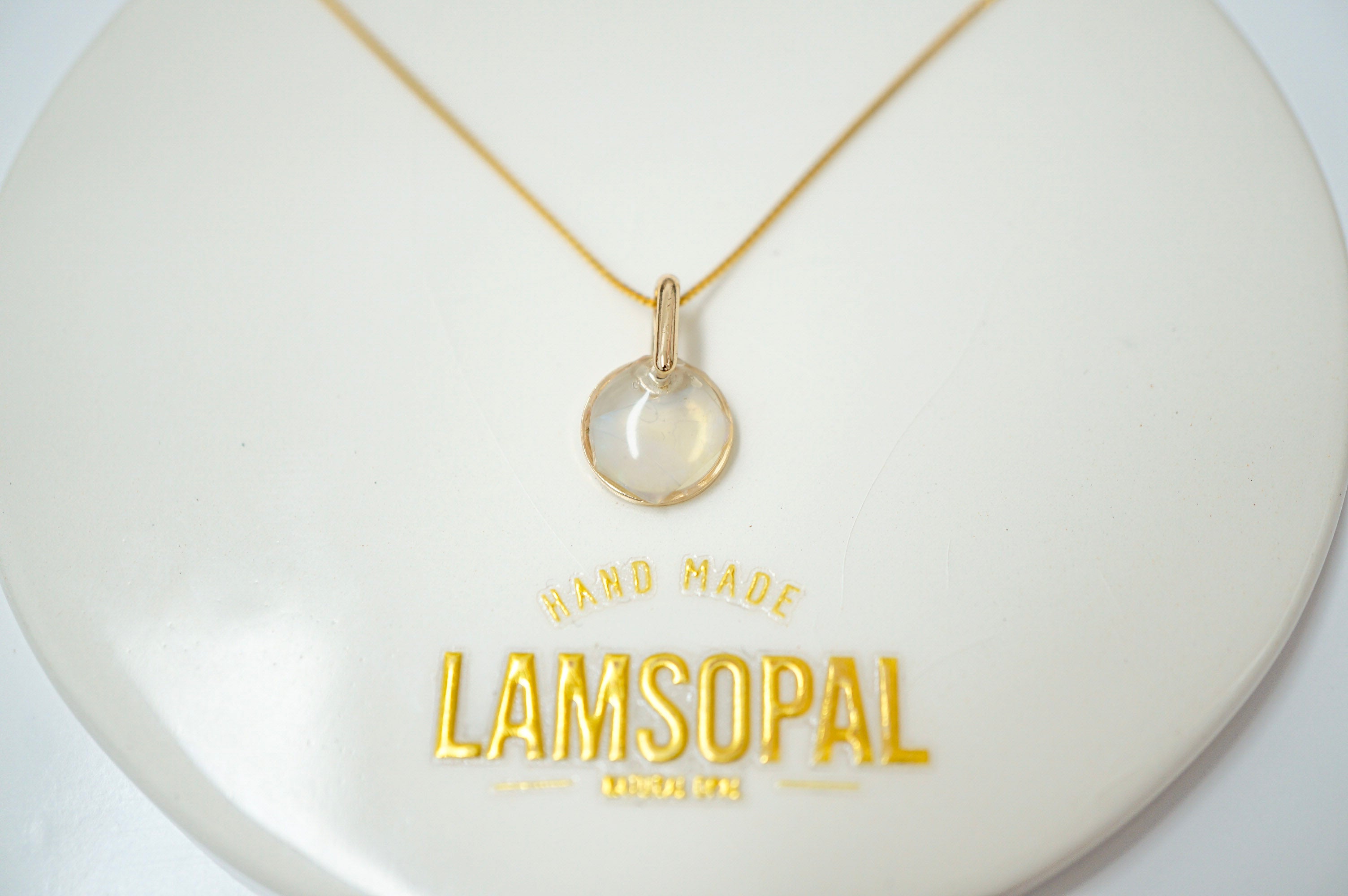 A delicate gold necklace with a small, round, clear cabochon pendant displayed on a white circular jewelry stand. The stand features gold foil text reading 'HAND MADE LAMSOPAL MUMBA'.