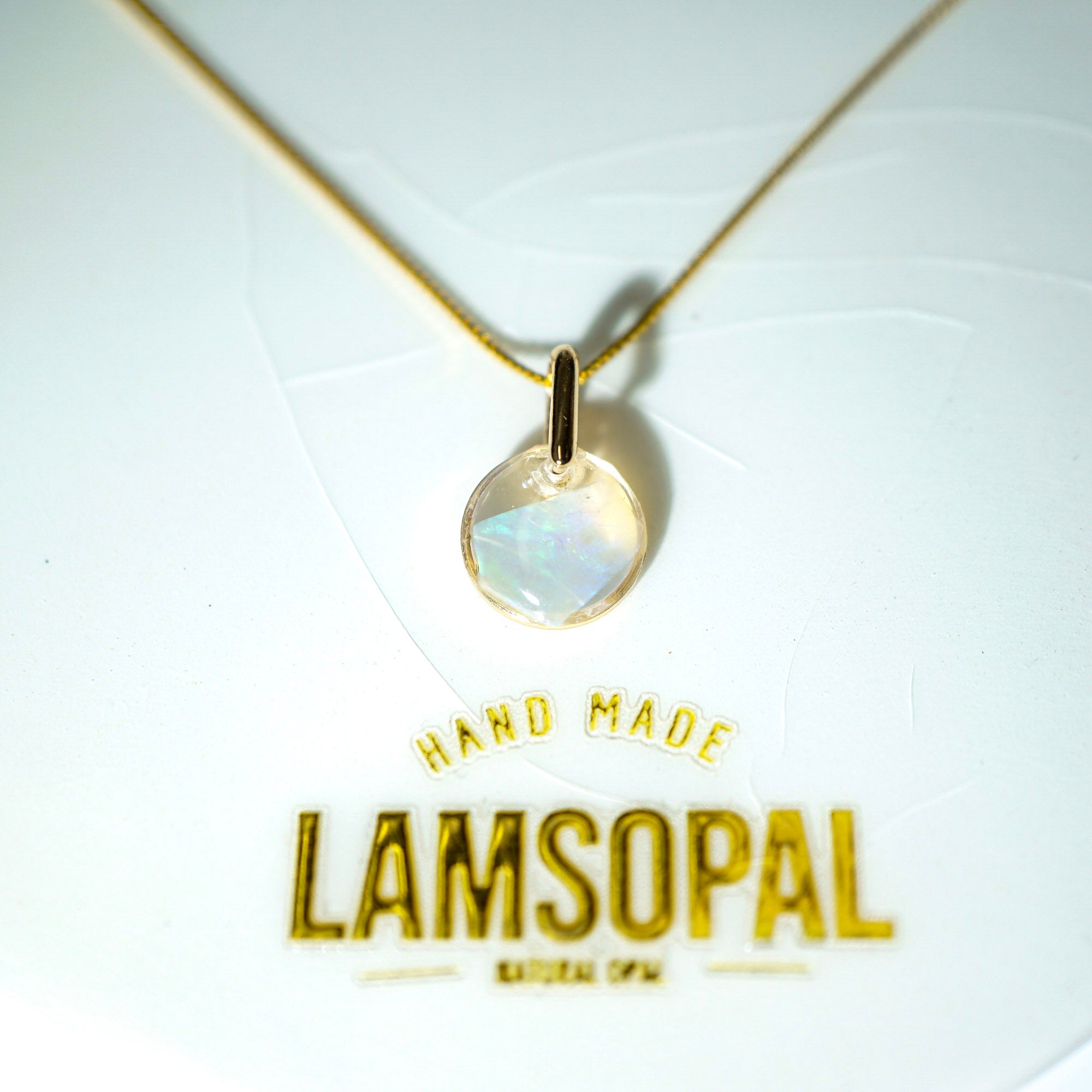 A gold chain necklace with a round iridescent moonstone pendant, displayed on a white background with gold foil branding that reads 'HAND MADE LAMSOPAL'.