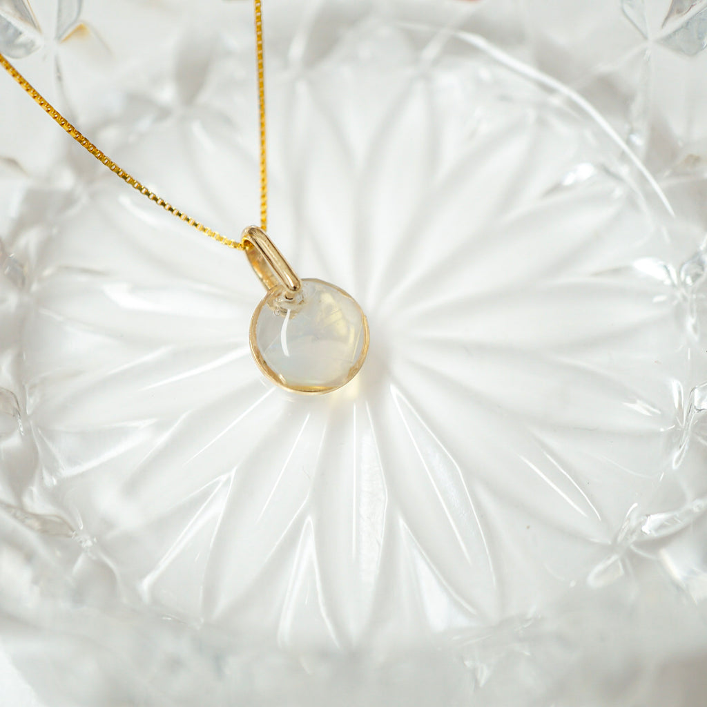 A gold chain necklace with a round, polished pale greenish-white pendant, displayed on a textured white glass plate.