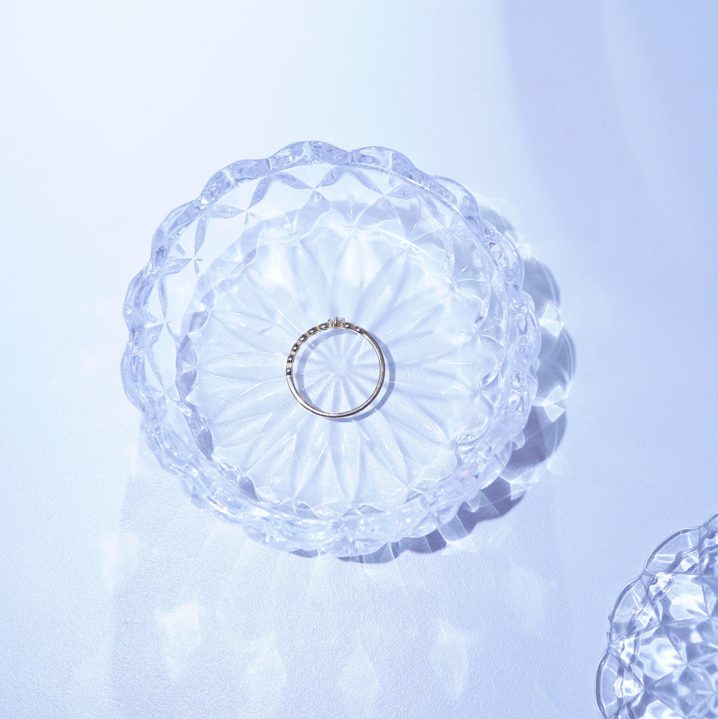 A delicate gold band ring with small clear stones, displayed inside a clear, faceted glass bowl on a light blue-white background.
