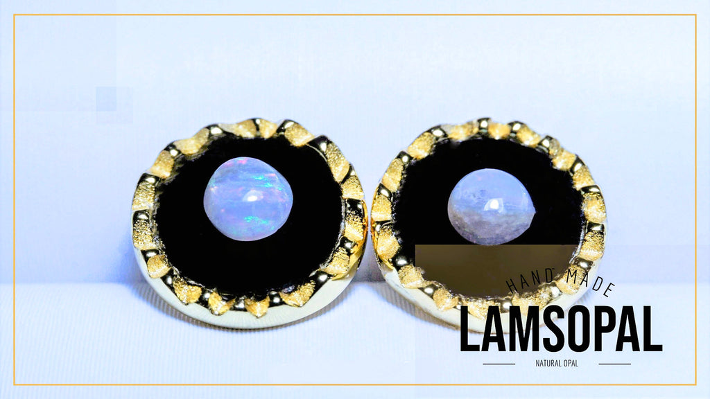 A pair of handmade Lamsopal stud earrings featuring oval natural opal centers set in black and gold bezel settings, displayed on a light blue background with a gold border.