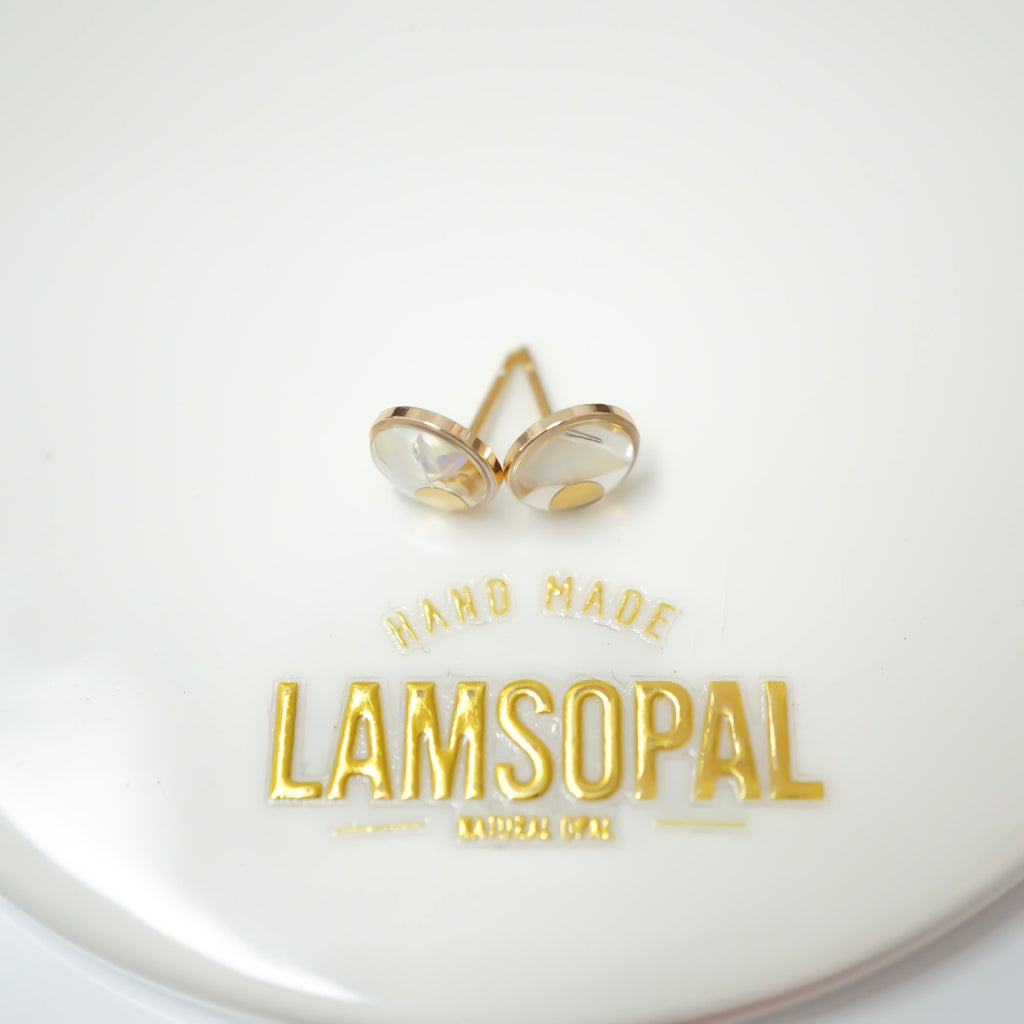A pair of gold-toned stud earrings with oval clear faceted stones, displayed on a white surface above gold foil text reading 'HAND MADE LAMSOPAL MINNE POL'.