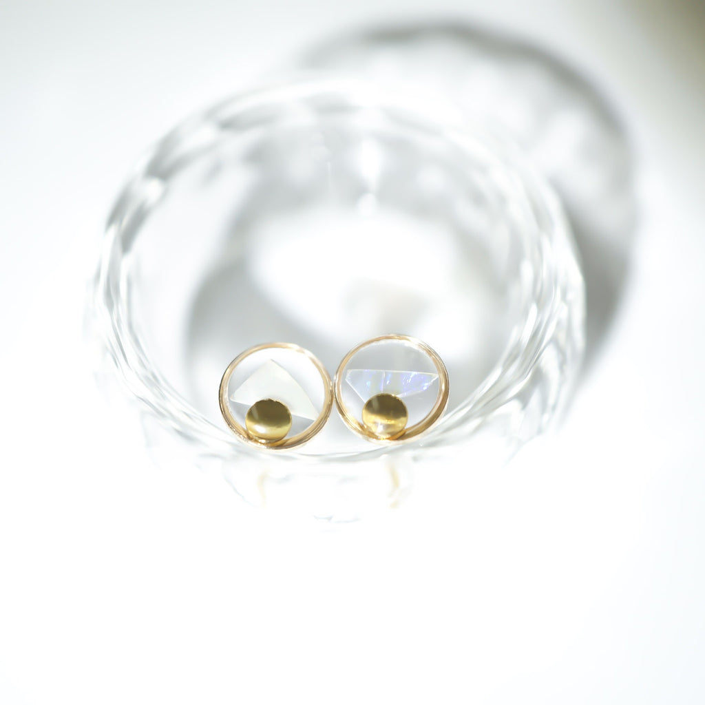 A pair of gold hoop earrings with faceted clear stones, displayed inside a textured clear glass dish on a bright white background.