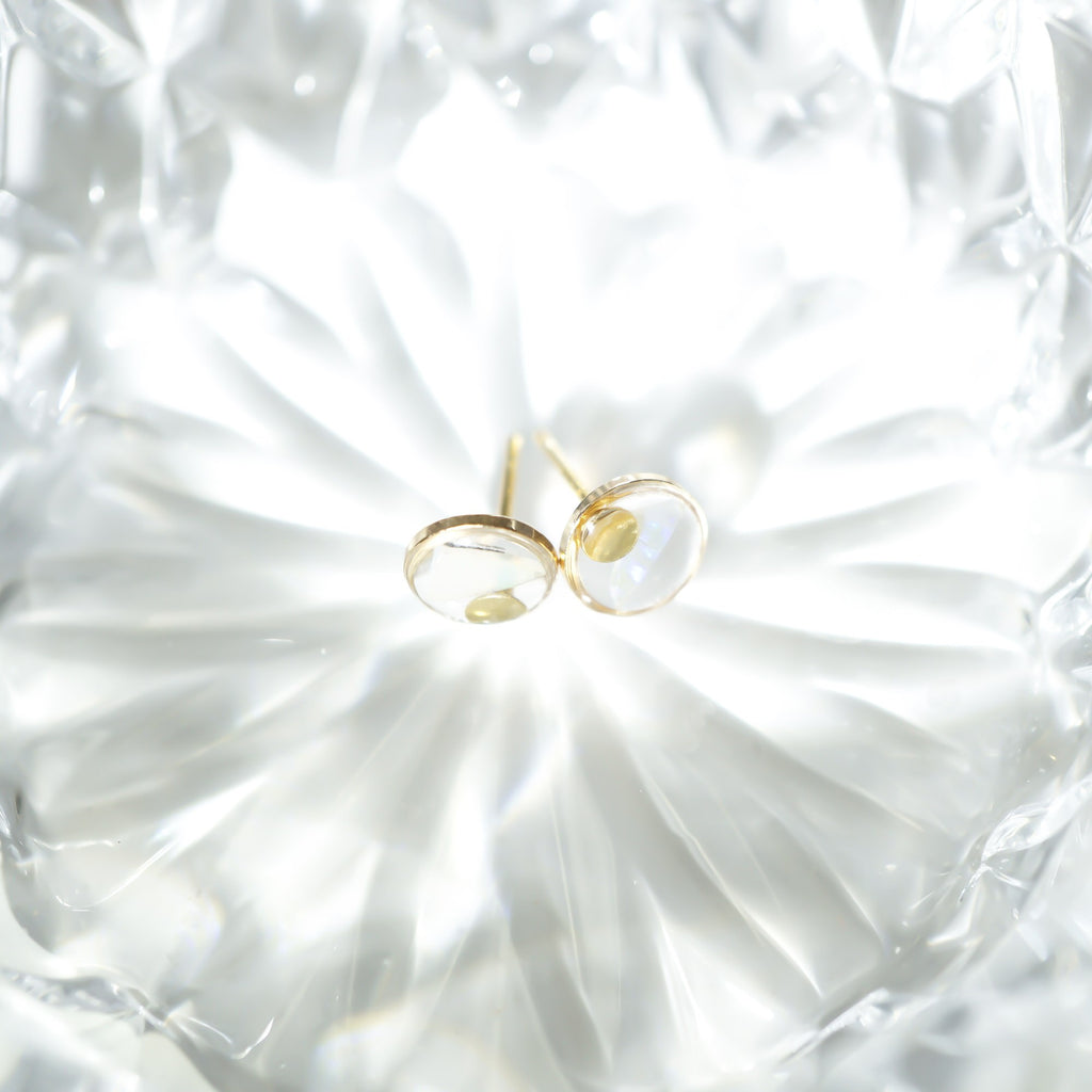 A pair of gold stud earrings with oval clear gemstones, resting on a textured, faceted white glass dish.