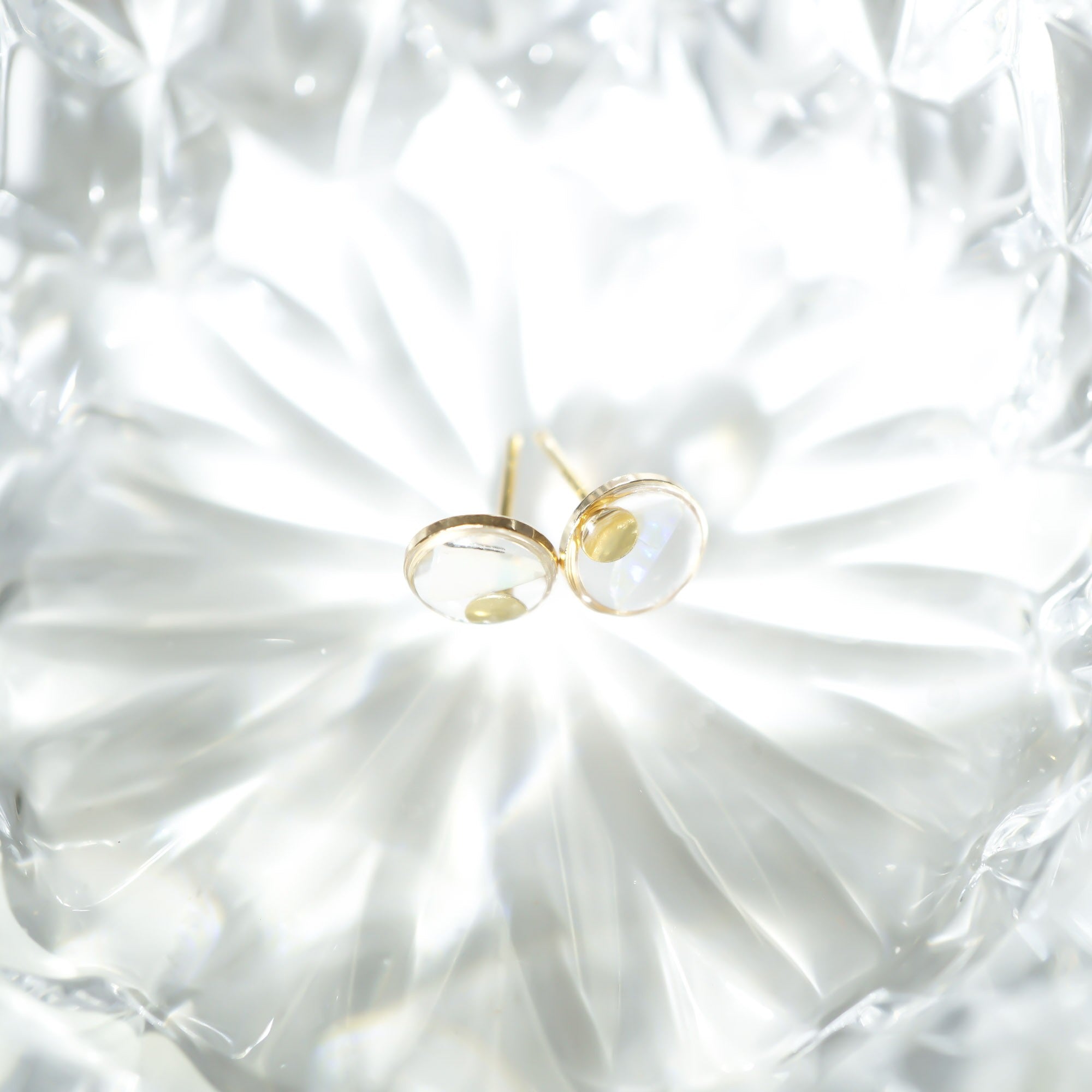 A pair of gold stud earrings with oval clear gemstones, resting on a textured, faceted white glass dish.