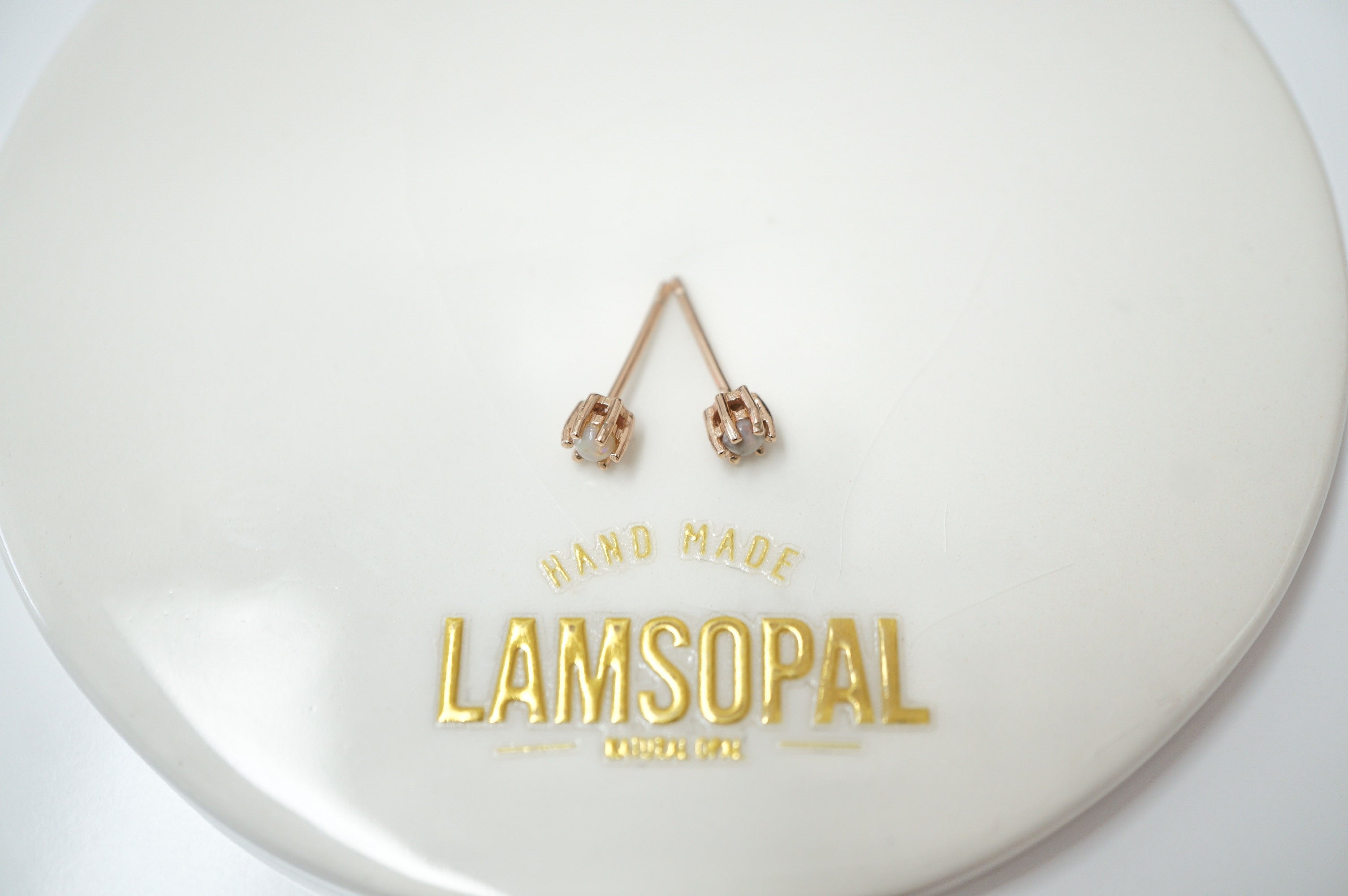 A pair of rose gold stud earrings with small clear stones, displayed on a white circular plate with gold foil branding that reads 'HAND MADE LAMSOPAL'.