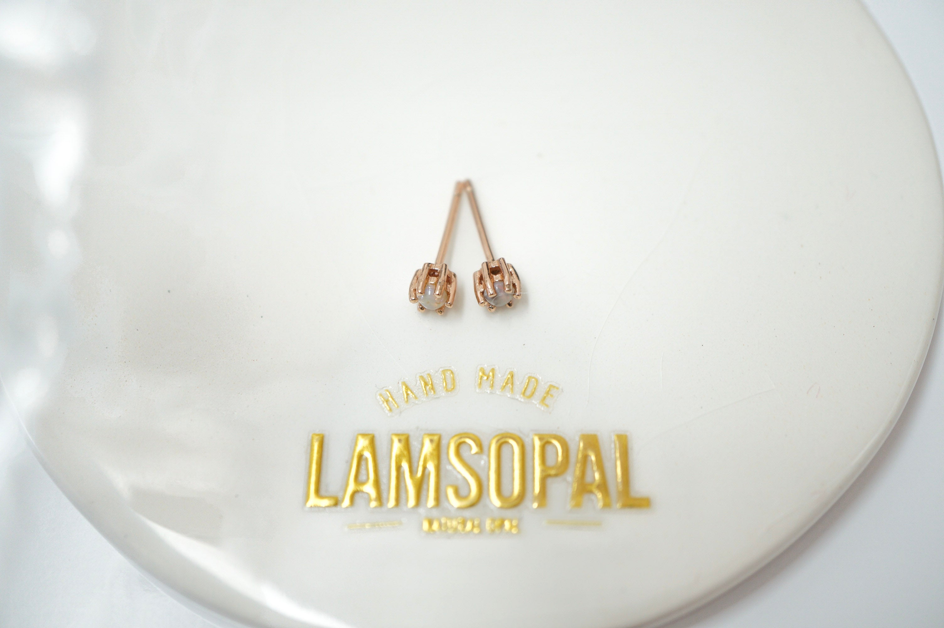 A pair of rose gold hoop earrings with small clear gemstone accents, displayed on a white circular stand with gold 'LAMSOPAL' branding.