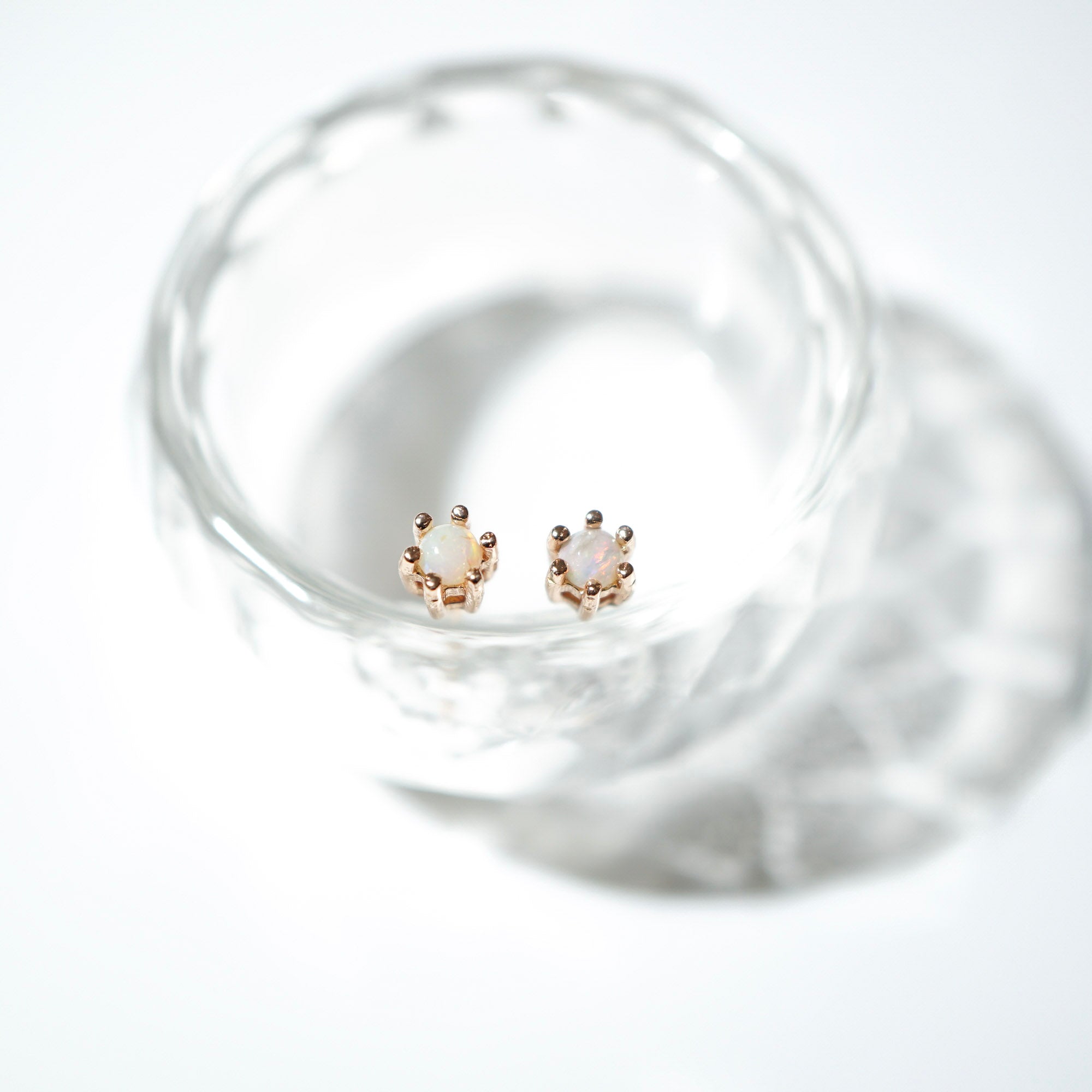 A pair of rose gold stud earrings with oval milky white gemstones, displayed inside a clear glass bowl on a bright white background.