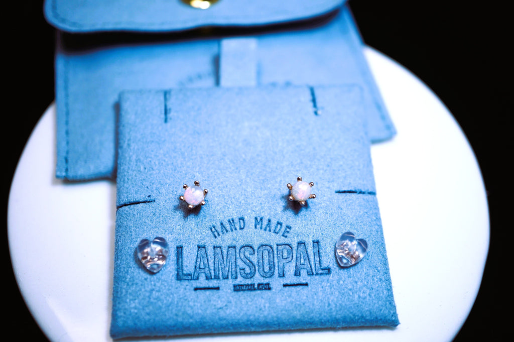 A pair of small stud earrings displayed on a blue felt jewelry card that reads 'HAND MADE LAMSOPAL', with matching blue pouches behind it on a white round surface against a black background.