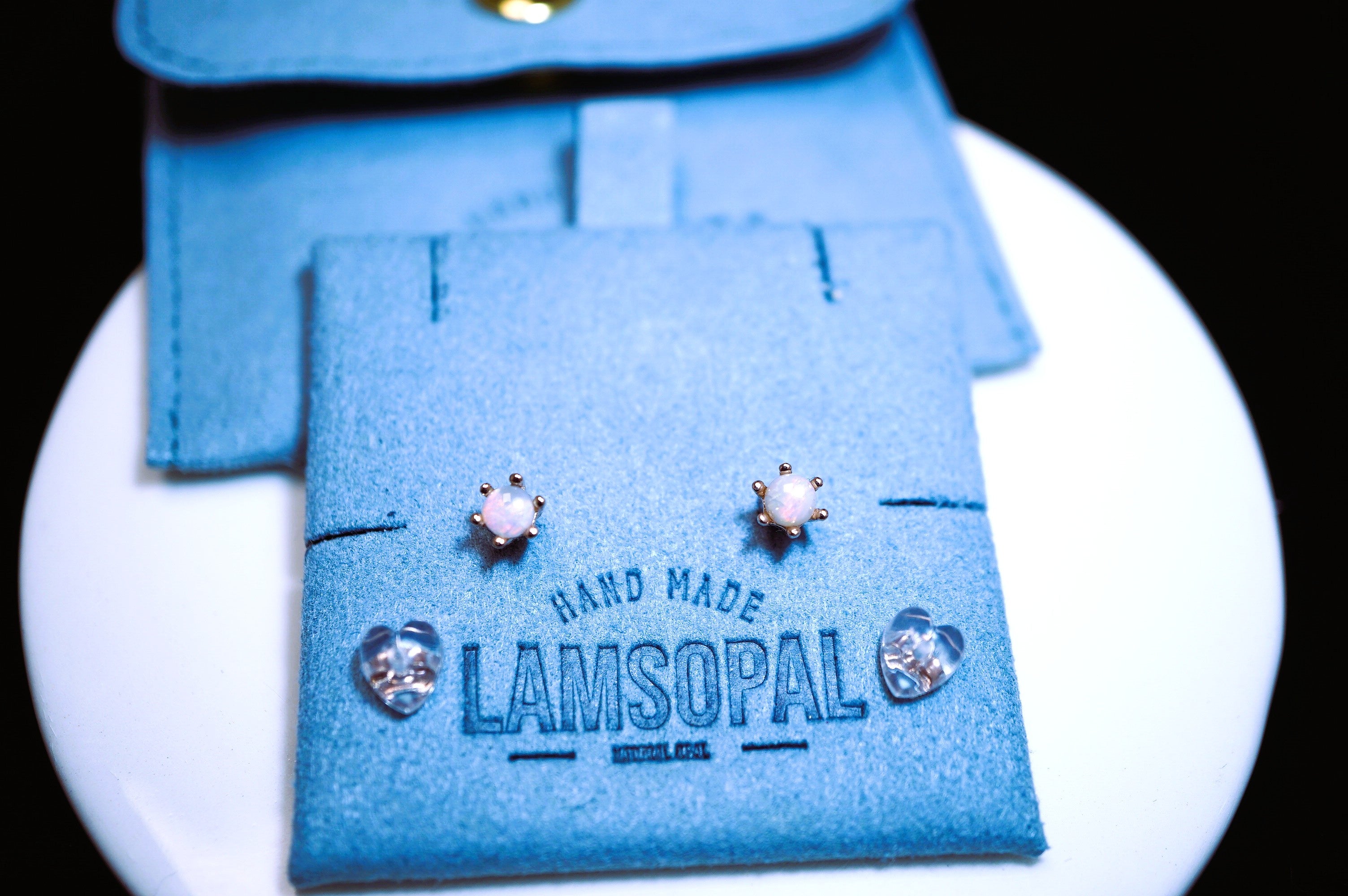 A pair of small stud earrings displayed on a blue felt jewelry card that reads 'HAND MADE LAMSOPAL', with matching blue pouches behind it on a white round surface against a black background.