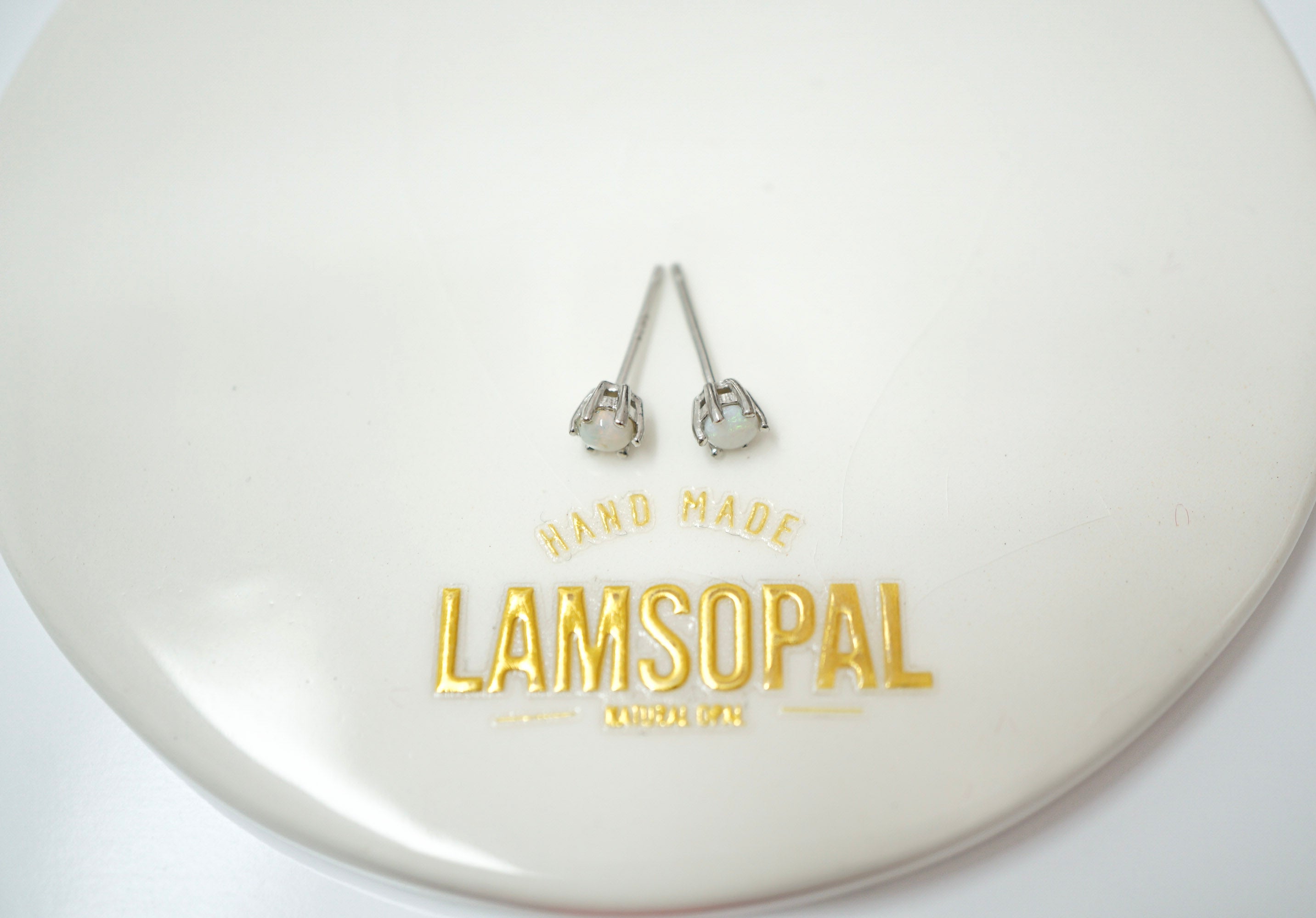 A pair of silver-toned stud earrings with clear heart-shaped stones, displayed on a glossy white circular plate with gold foil branding that reads 'HAND MADE LAMSOPAL'.