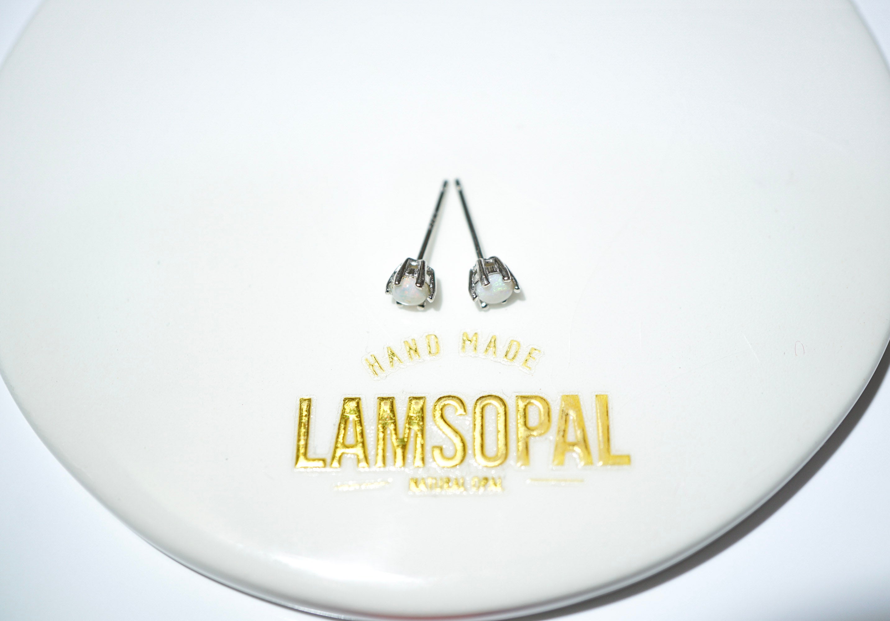A pair of silver stud earrings with clear faceted gemstones, displayed on a white circular plate with gold foil branding that reads 'HAND MADE LAMSOPAL'.
