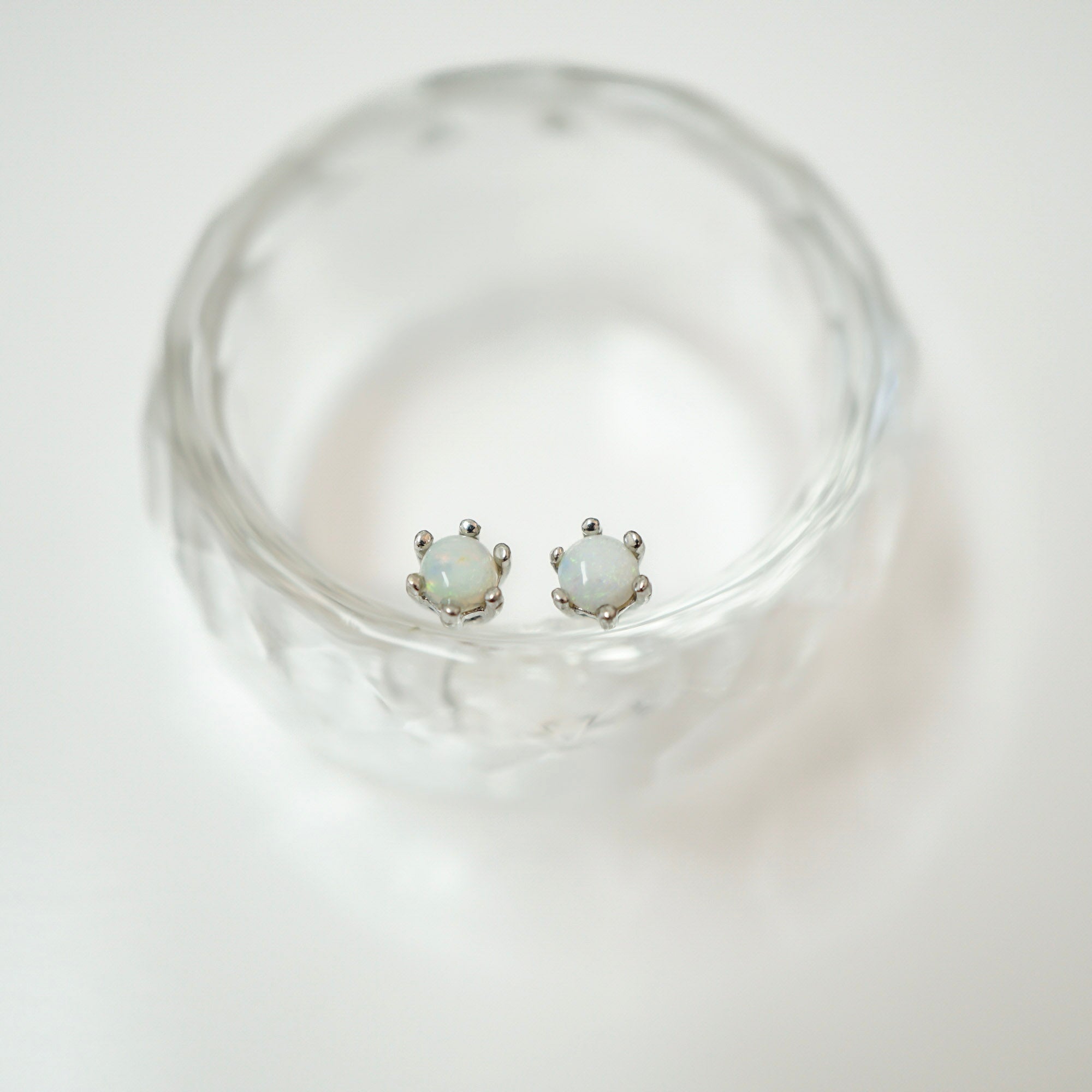 A pair of silver stud earrings with oval iridescent white stones, displayed inside a clear textured glass bowl on a white background.