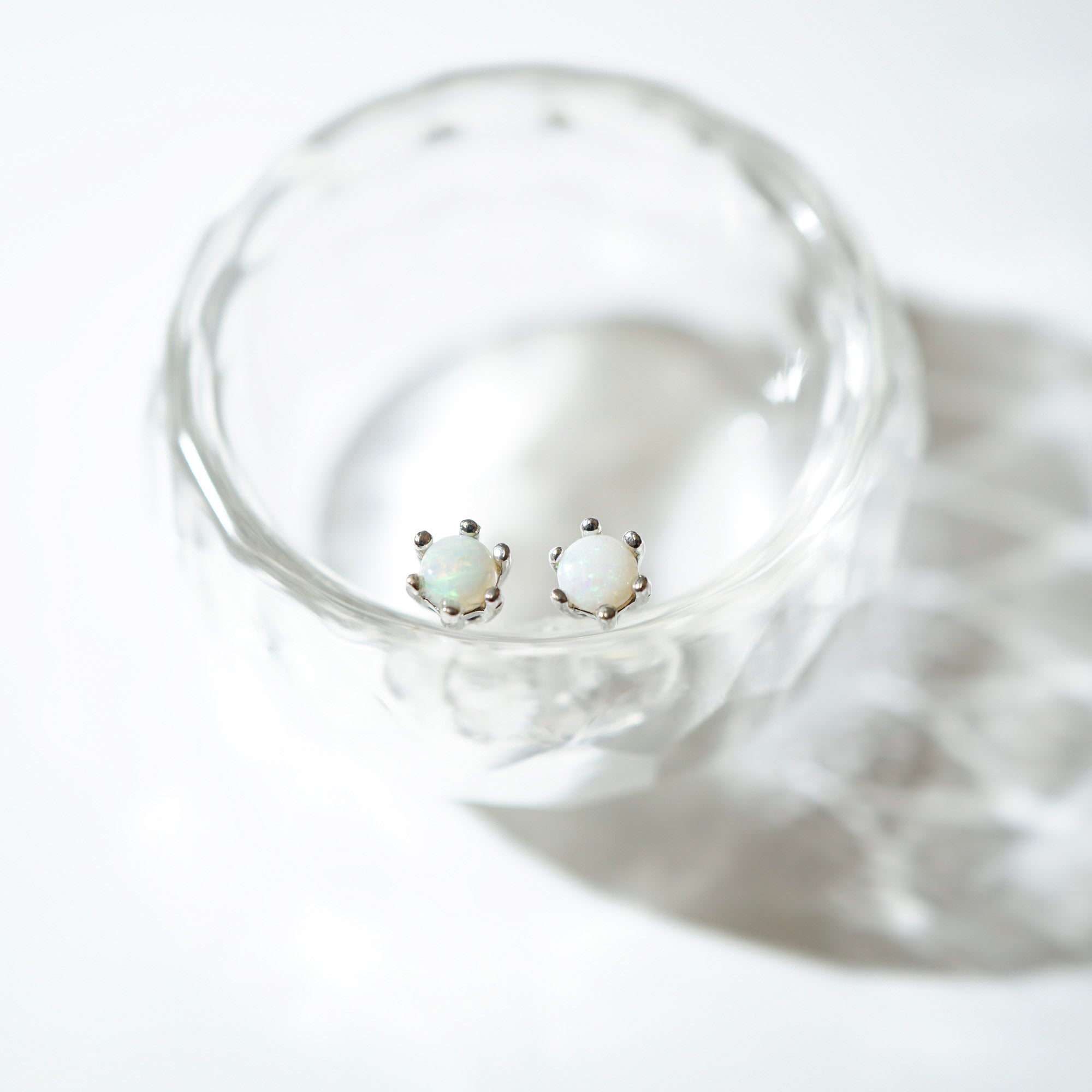 A pair of small stud earrings with round white opal-like centers in silver settings, displayed inside a clear glass dish on a bright white background.