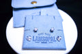 A pair of small stud earrings displayed on a blue felt jewelry card that reads 'HAND MADE LAMSOPAL', with a matching blue felt pouch behind it on a white round surface against a black background.
