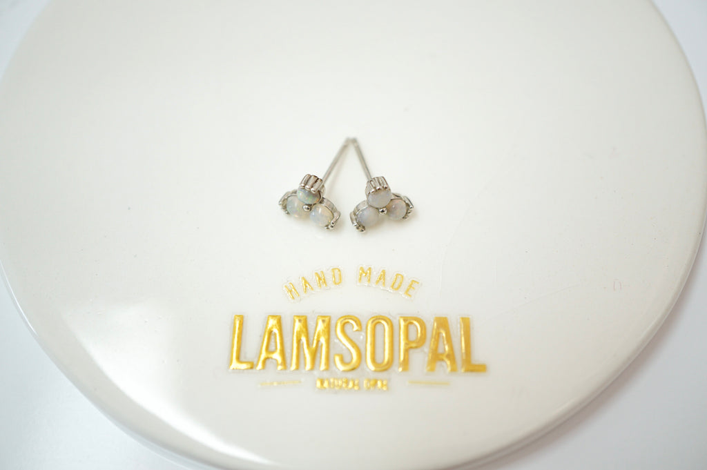 A pair of silver-toned stud earrings with iridescent white stones, displayed on a glossy white circular surface printed with the brand name 'LAMSOPAL'.
