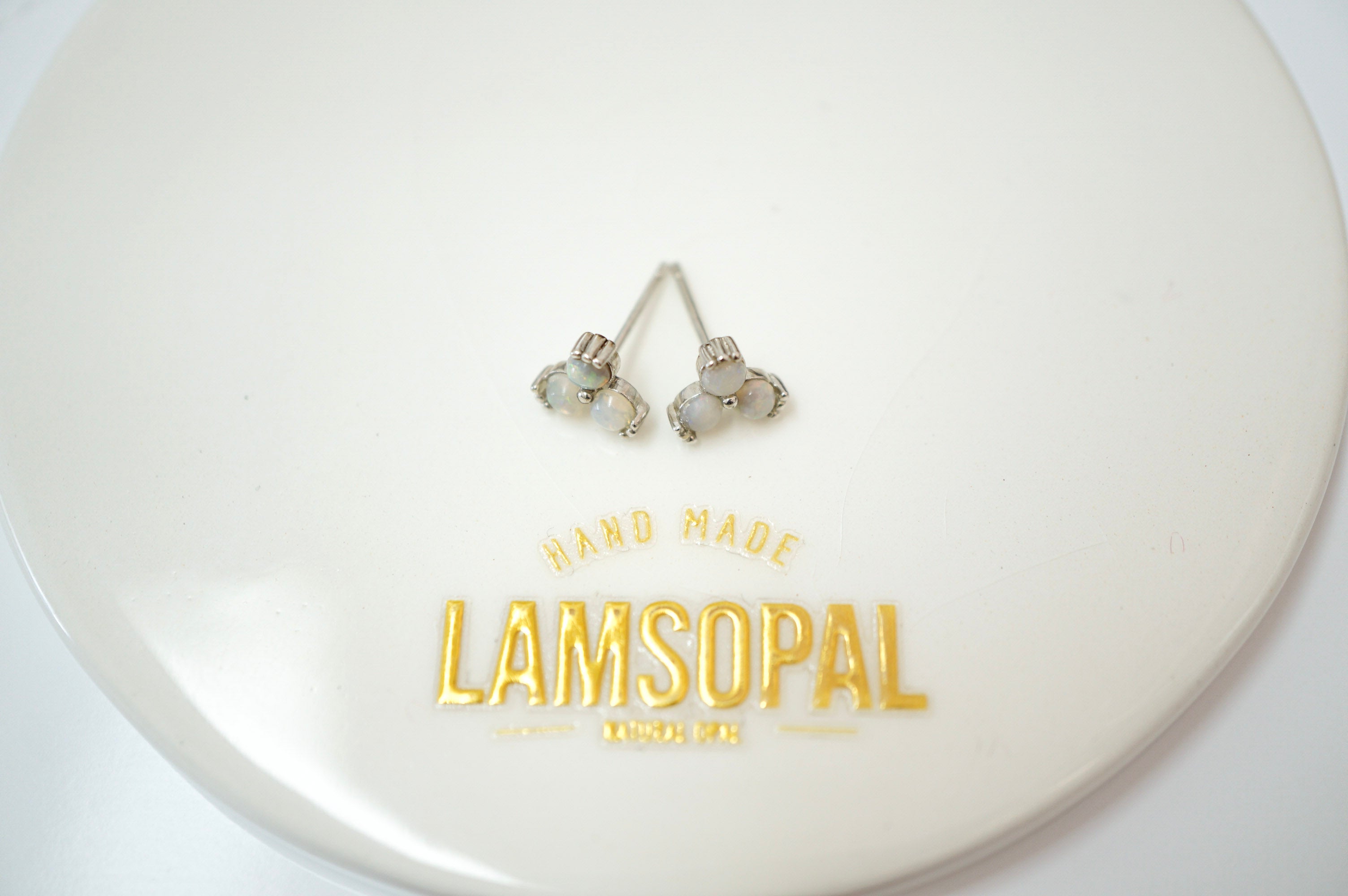 A pair of silver-toned stud earrings with iridescent white stones, displayed on a glossy white circular surface printed with the brand name 'LAMSOPAL'.