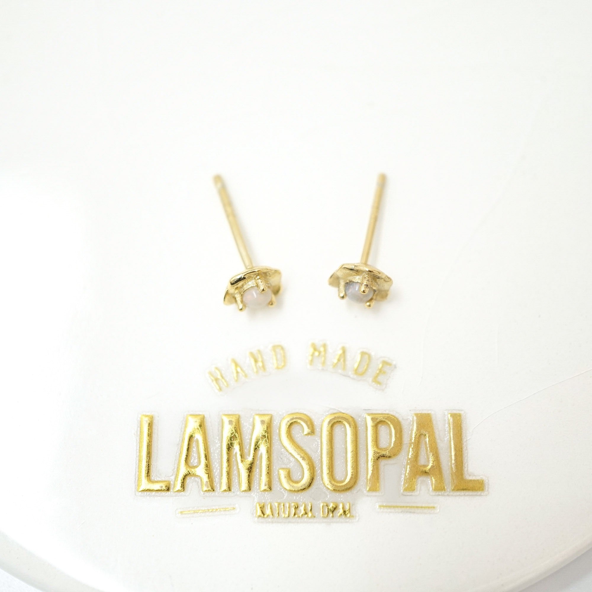 A pair of small gold stud earrings with clear gemstones in a prong setting, displayed on a white background above a gold 'LAMSOPAL' brand logo and 'HAND MADE' text.