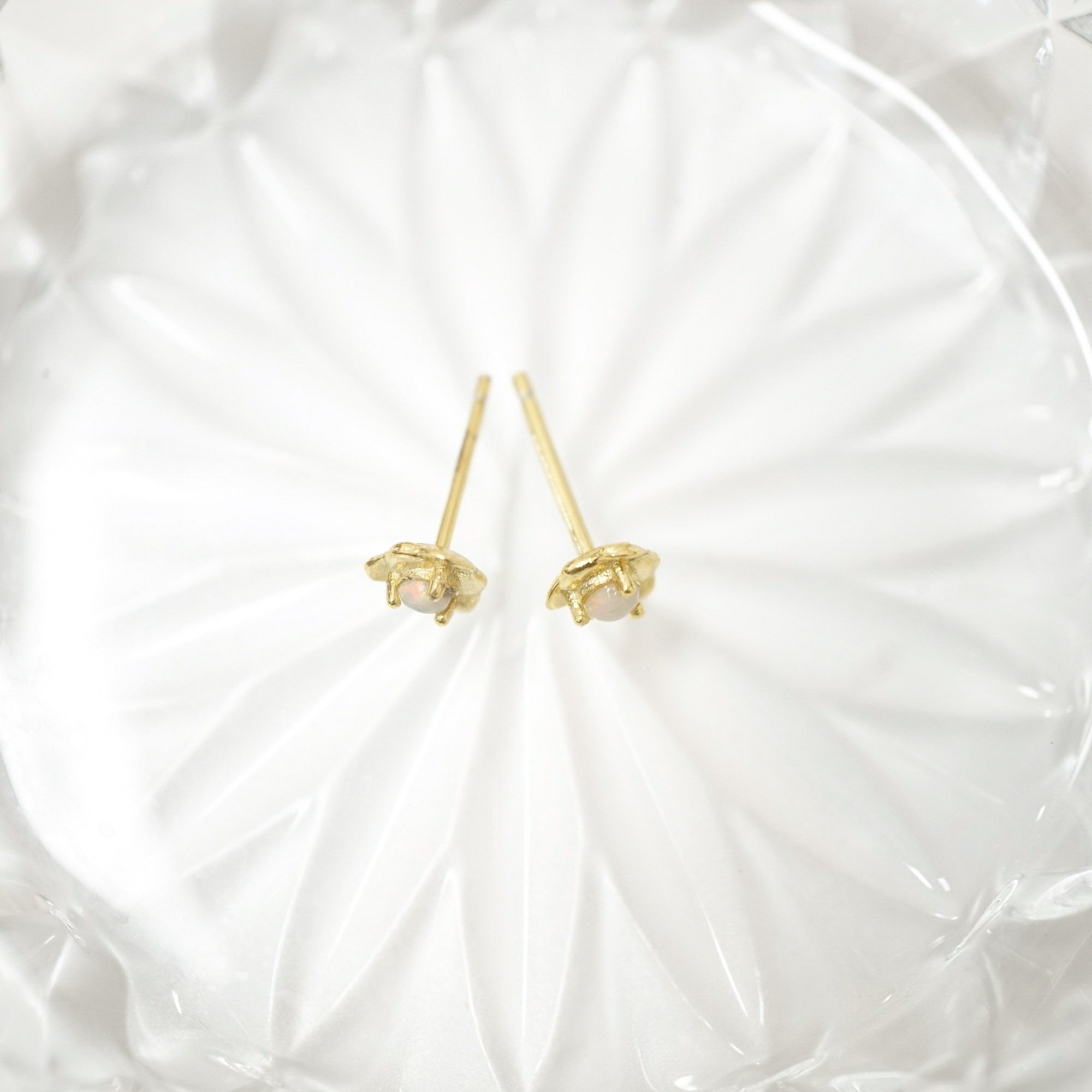 A pair of small gold stud earrings with pale pink stones, displayed on a white textured glass dish.
