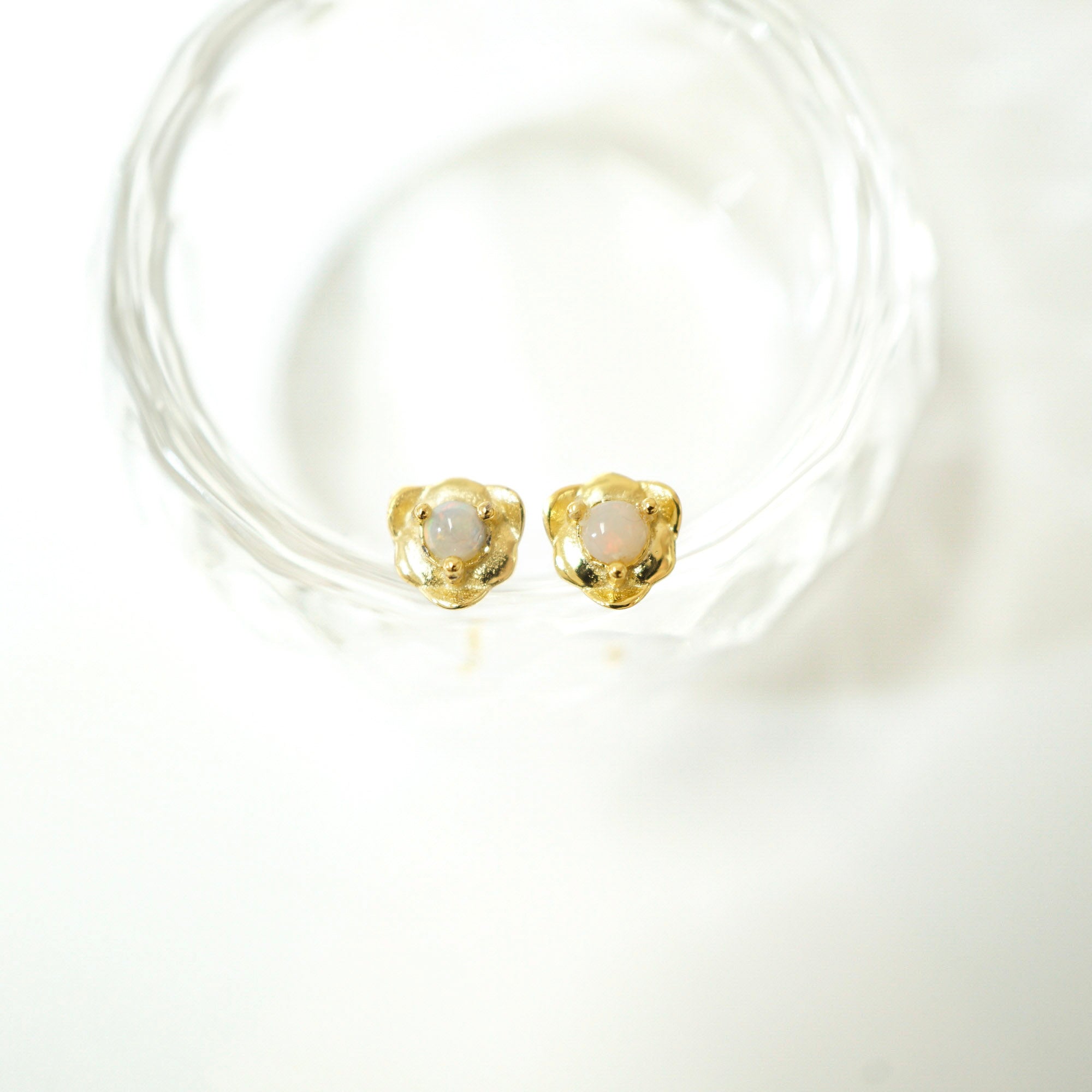 A pair of gold-toned stud earrings with oval milky white stones, displayed on a clear textured glass coaster against a bright white background.