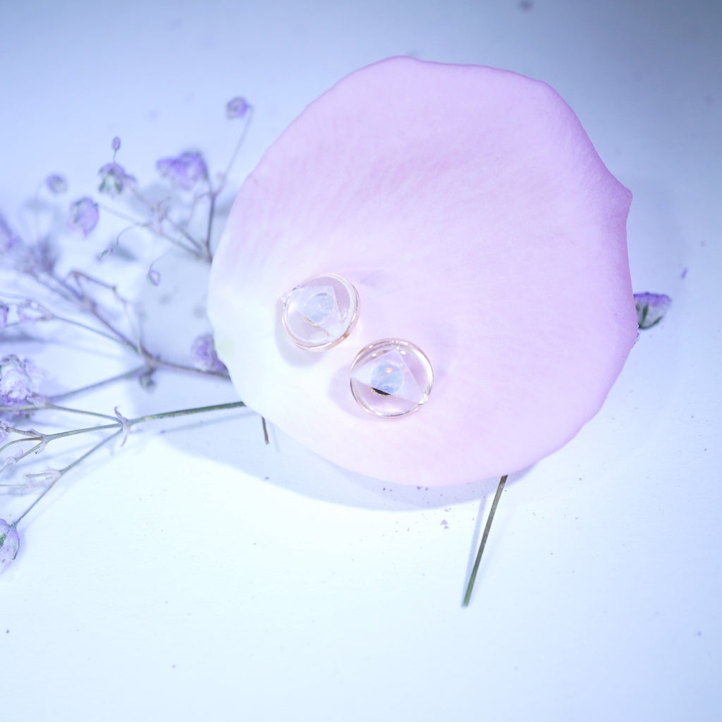 Two clear oval gemstones with a soft pink iridescent sheen, resting on a light pink rose petal with dried lavender flowers on a pale blue-white background.