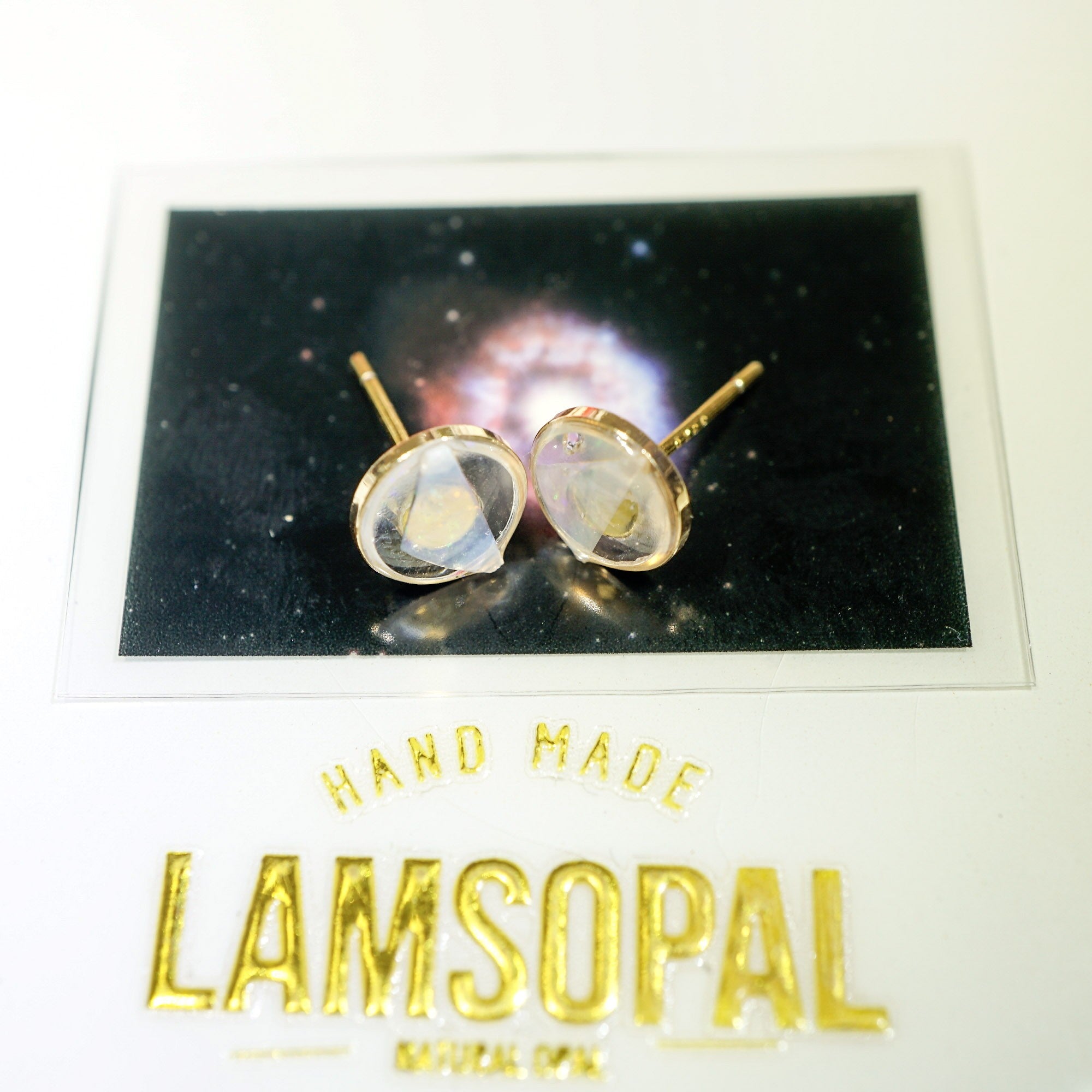 A pair of gold-toned stud earrings with oval clear stones, displayed on a branded card that reads 'HAND MADE LAMSOPAL'.