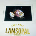 A pair of gold stud earrings with oval iridescent cabochon gemstones displayed on a black card, mounted on a white backing card with gold text reading 'HAND MADE LAMSOPAL NATURAL OPAL'.