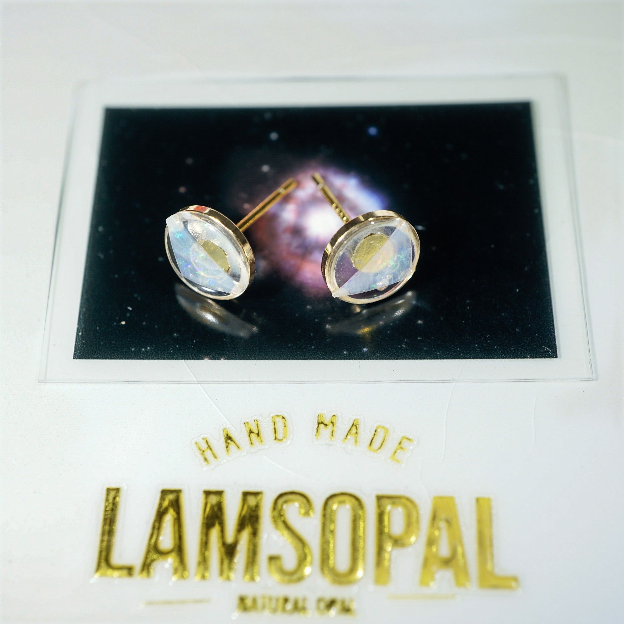 A pair of gold stud earrings with oval iridescent cabochon gemstones displayed on a black card, mounted on a white backing card with gold text reading 'HAND MADE LAMSOPAL NATURAL OPAL'.