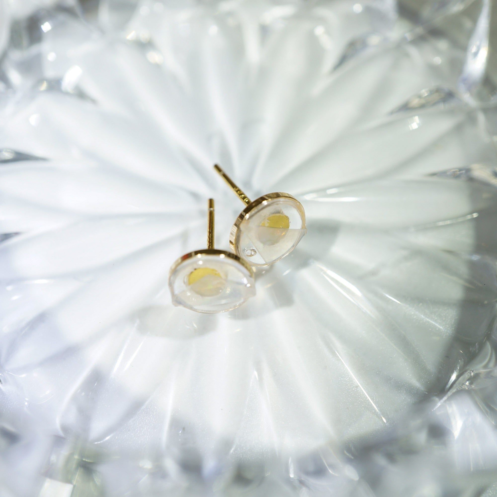 A pair of gold stud earrings with oval clear resin pendants containing tiny yellow inclusions, resting on a white textured surface.