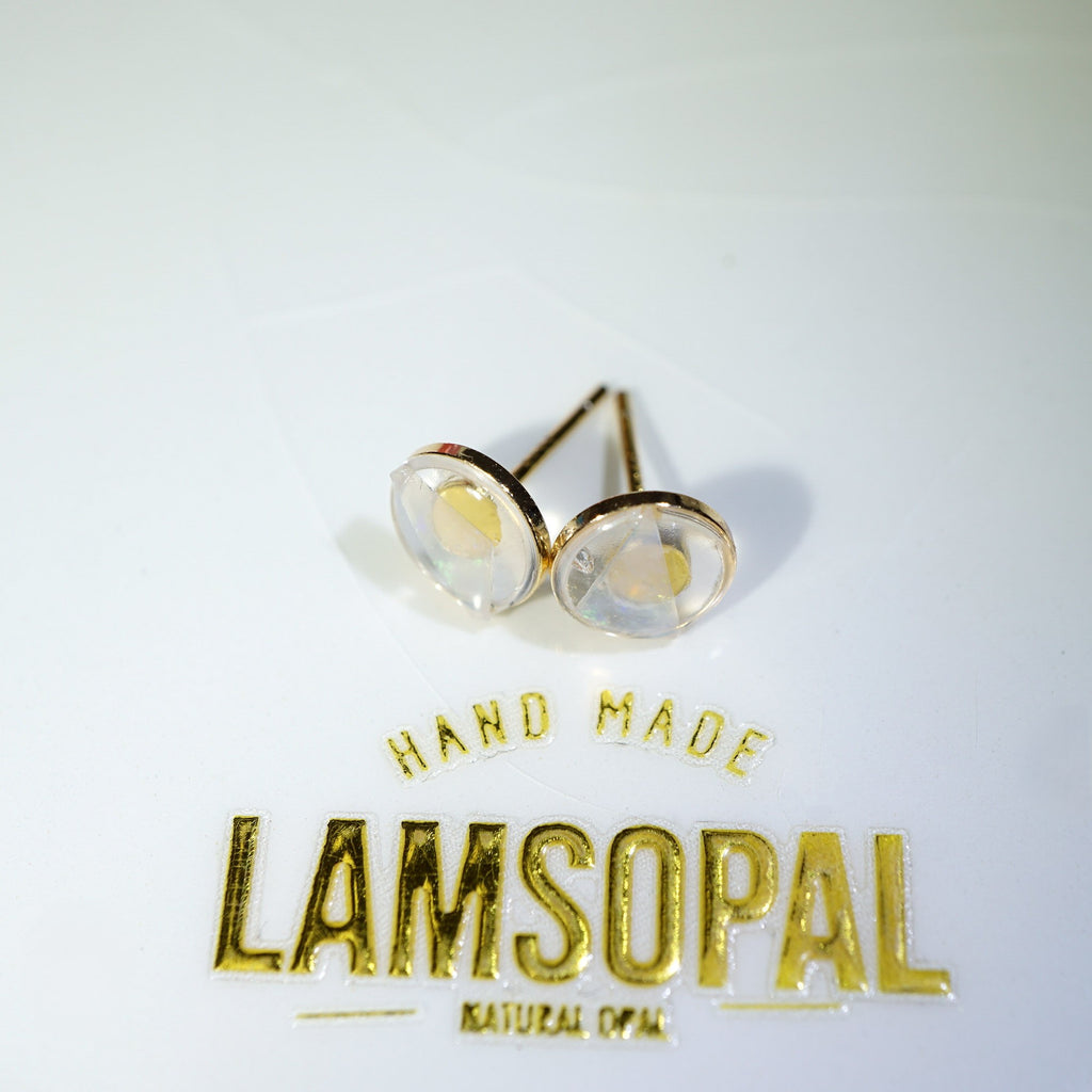 A pair of gold post stud earrings with round clear cabochon gemstones, displayed on a white surface above a gold foil brand label reading 'LAMSOPAL'.
