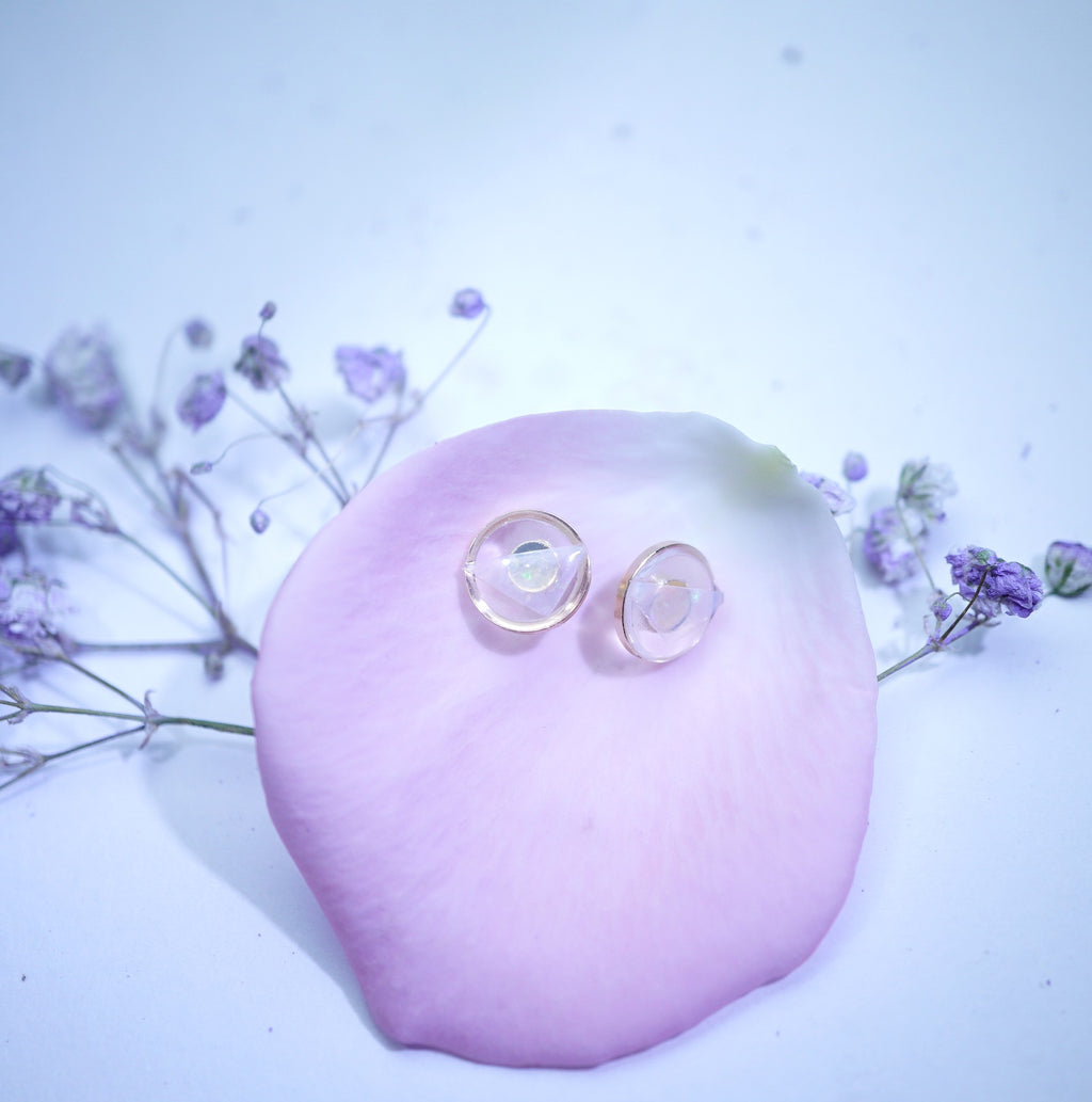 Two clear contact lenses with gold rims resting on a light pink rose petal, with small purple flowers in the background on a pale blue-white surface.
