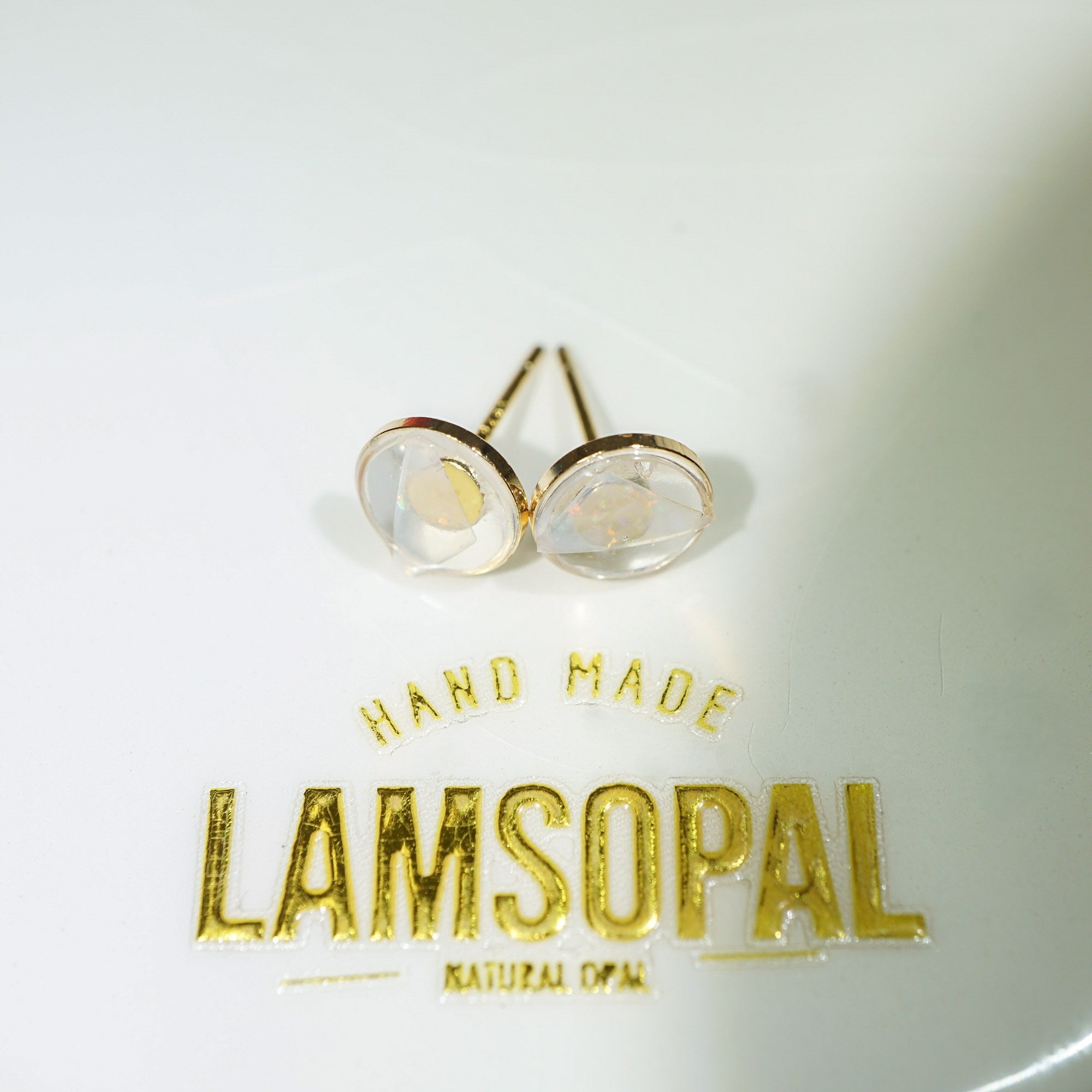 A pair of gold stud earrings with oval clear resin pendants, displayed on a white surface above gold foil text reading 'HAND MADE LAMSOPAL'.