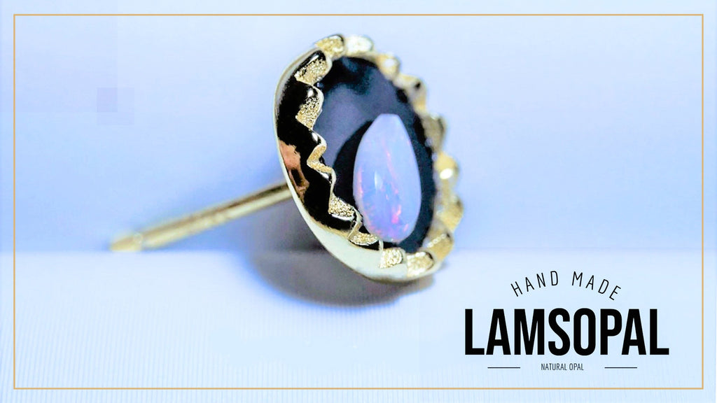 A gold-toned Lamsopal stud earring with a natural opal gemstone showing blue and purple iridescence, set against a light blue background with a gold border. The text 'HAND MADE LAMSOPAL' is visible in the bottom right corner.