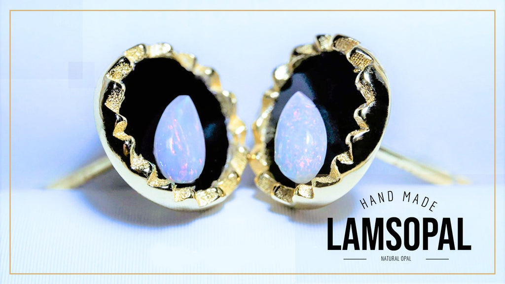 A pair of handmade Lamsopal stud earrings featuring natural opal stones with a blue iridescent sheen, set in gold bezel settings with a black background and textured gold borders. The image includes a text overlay on the bottom right reading 'HAND MADE LAMSOPAL NATURAL OPAL'.