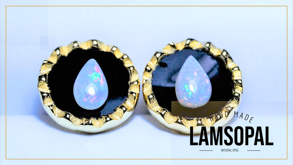 A pair of Lamsopal natural opal teardrop stud earrings set in gold and black bezel settings, displayed on a light blue background with a gold border. The text 'LAMSOPAL' is visible in the bottom right corner.