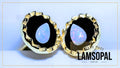 A pair of Lamsopal handmade stud earrings featuring teardrop-shaped natural opal stones with a blue and pink iridescent play-of-color, set in textured gold bezels with a black center. The image is presented on a light blue background with a thin gold border and a brand logo overlay in the bottom right corner.